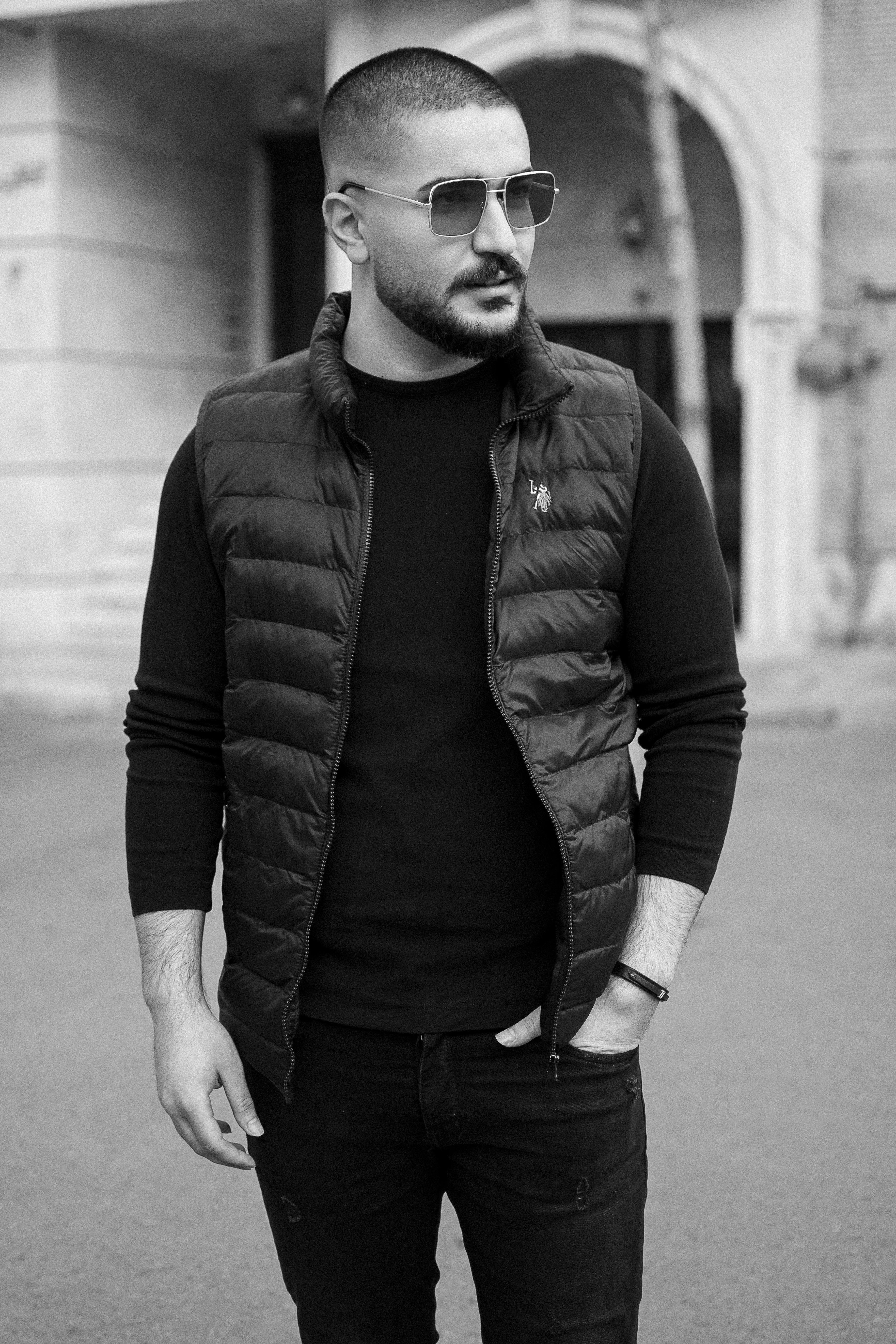 man in black jacket and black pants standing on sidewalk in grayscale photography